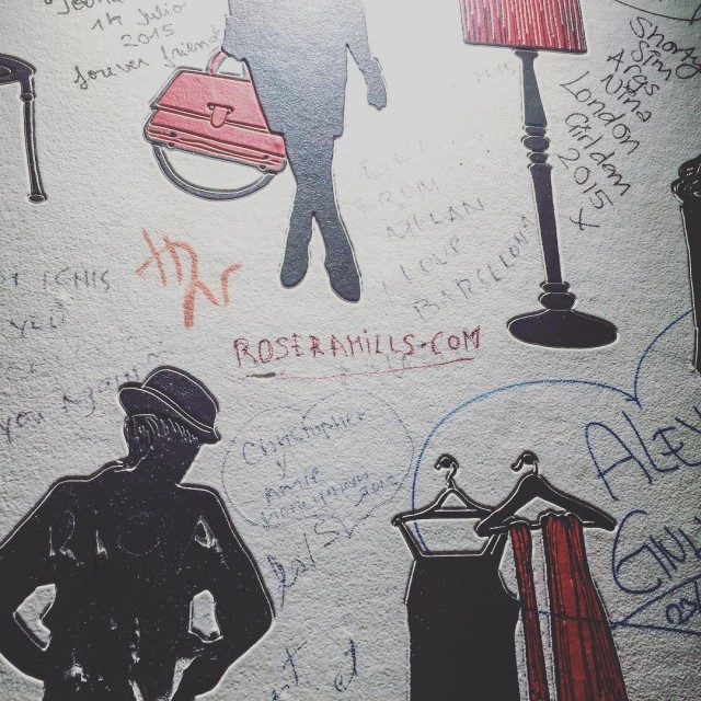 Pared blanca con dibujos de un bolso, una lámpara y perchas, llena de firmas de visitantes, con roseramills.com escrito en rojo