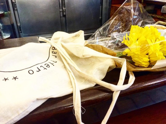 Bolsa de tela del Bar Pietro junto a un ramo de margaritas amarillas sobre una barra