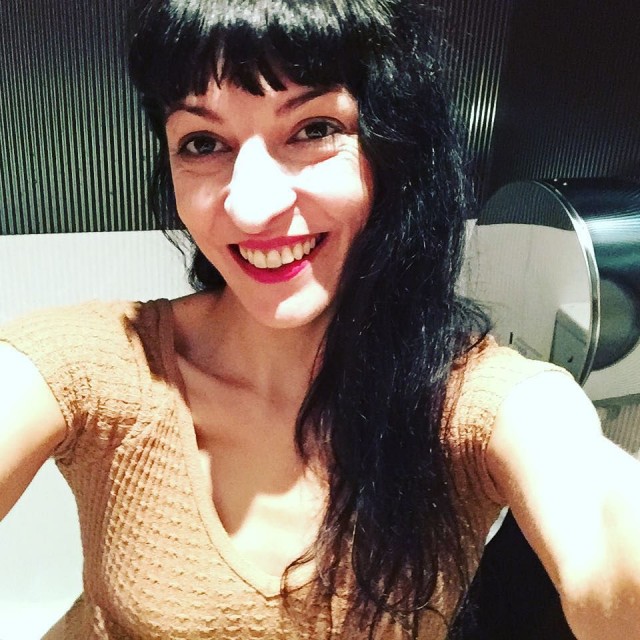 Roser Amills sonríe con los labios rojos en un selfie en un baño público, con camiseta de punto beige y un dispensador de papel al fondo