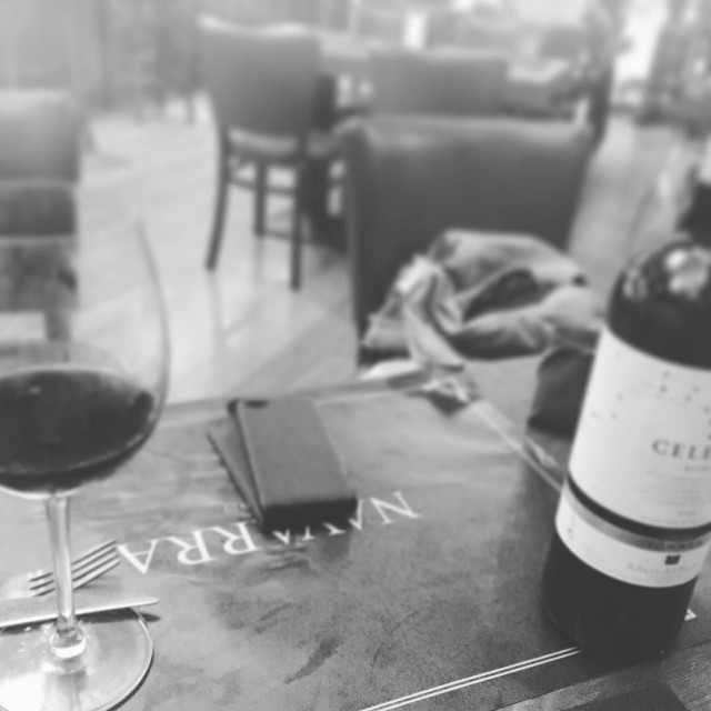 Copa de vino tinto, un móvil y una botella de vino sobre una mesa de bar con un salvamanteles donde se lee Navarra, en blanco y negro
