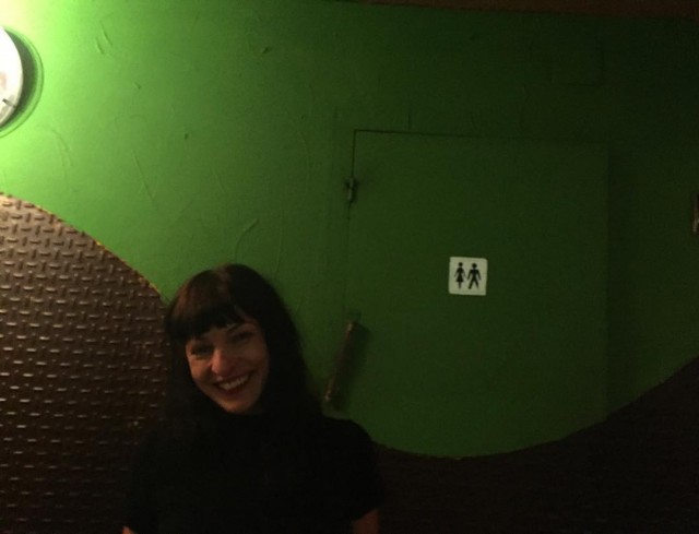Roser Amills sonriendo ante una pared verde con la puerta de los lavabos, dentro de El Otro Bar