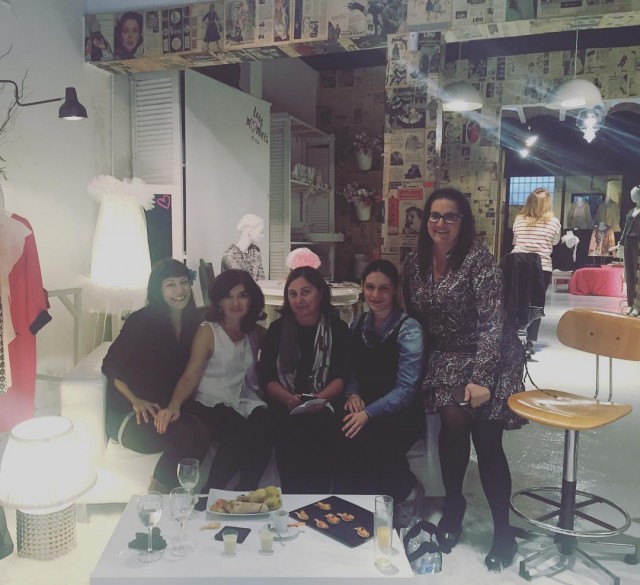Cinco mujeres posan, sentadas y de pie, junto a una mesa con copas y aperitivos, en una sala con paredes forradas de recortes de revistas y prendas sobre maniquíes