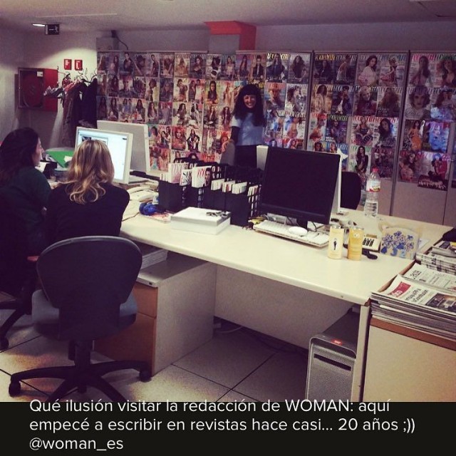 Redacción de la revista Woman, con una pared de portadas de revistas al fondo, dos redactoras sentadas ante sus pantallas y una persona de pelo oscuro de pie al fondo