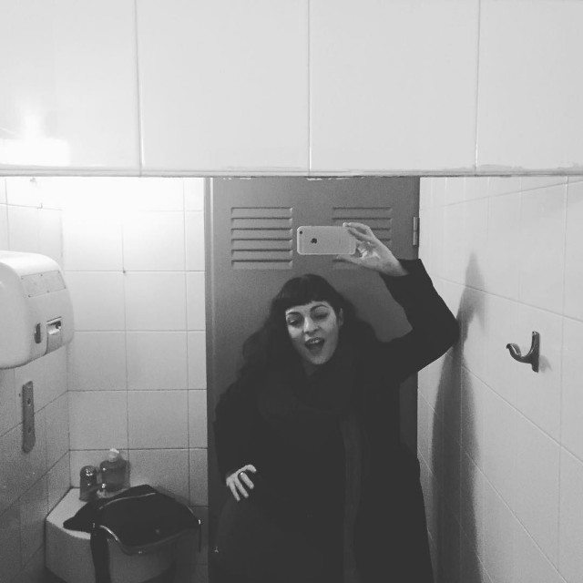 Selfi en blanco y negro ante el espejo de un cuarto de baño: una mujer con flequillo y abrigo oscuro, sonriendo, sostiene un móvil