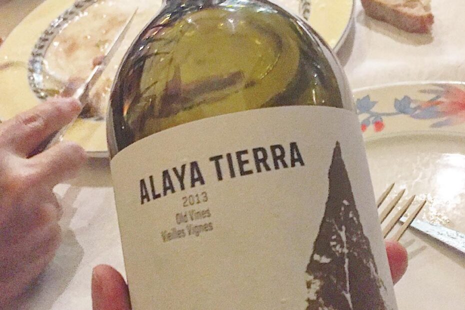 Mano sosteniendo una botella de vino Alaya Tierra 2013 Old Vines sobre una mesa con platos usados