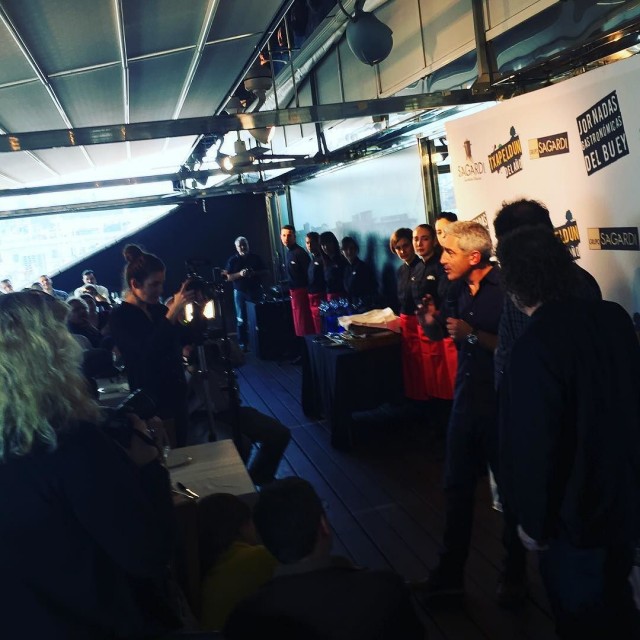Acto de las Jornadas Gastronómicas del Buey de Grupo Sagardi: un hombre canoso hablando de pie, rodeado de cocineros con delantal rojo, ante un panel con los logos de Txapeldun y Sagardi