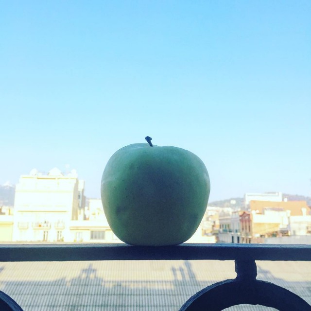 Una manzana verde sobre una barandilla de hierro, a contraluz, con edificios de Barcelona y un cielo azul claro al fondo