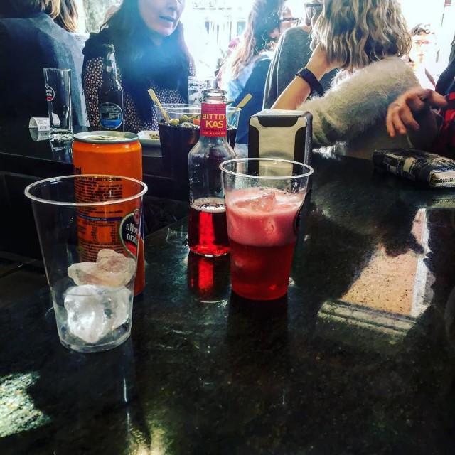Barra de bar con un vaso de plástico con hielo y bebida roja, una lata de naranjada y una botella de Bitter Kas, con gente al fondo