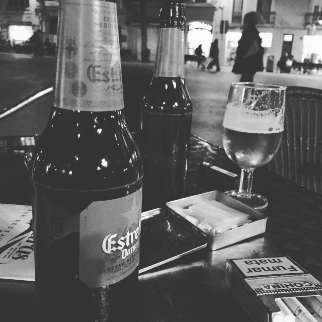 Fotografía en blanco y negro de dos botellas de cerveza Estrella Damm y una copa sobre la mesa de una terraza, junto a un paquete de tabaco