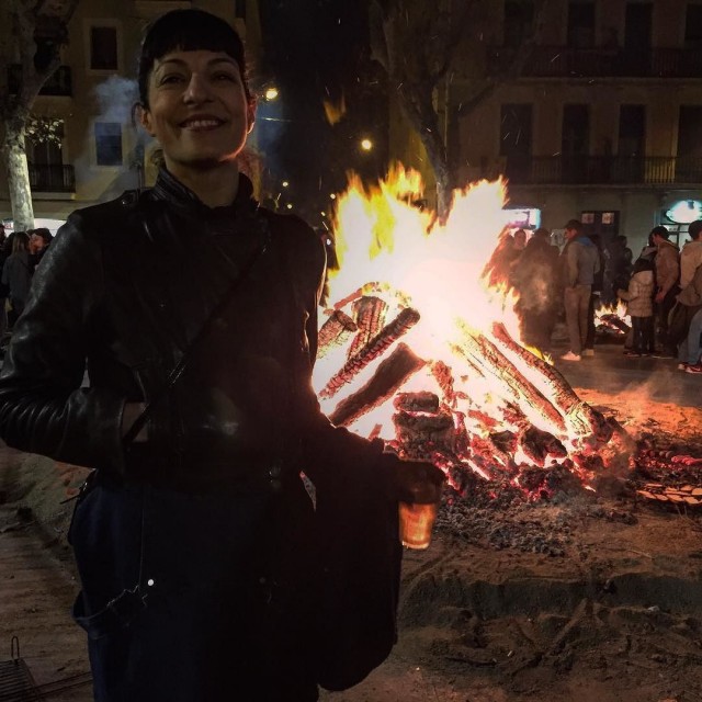 Persona somrient amb jaqueta negra davant d'un fogueró encès en una plaça de Gràcia