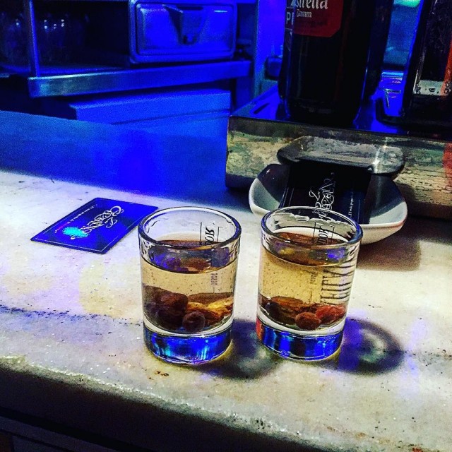 Dos chupitos con licor ámbar y fruta sobre la barra de un bar, con luz azul al fondo