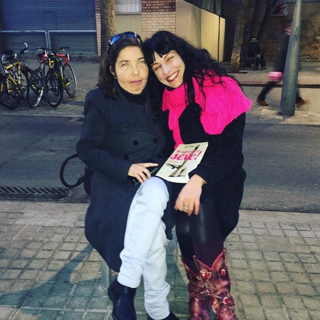 Dos mujeres abrazadas sentadas en un banco de la calle; una sostiene un libro con la palabra «sexe» en la portada