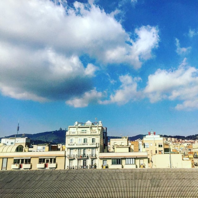 Nubes blancas en un cielo azul sobre azoteas y edificios de una ciudad, con montañas al fondo