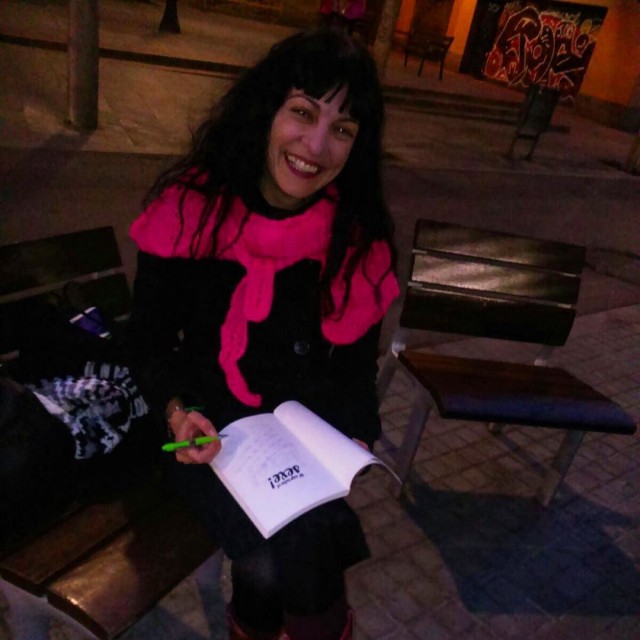 Dona somrient asseguda en un banc de nit, amb un llibre obert i un bolígraf verd a la mà
