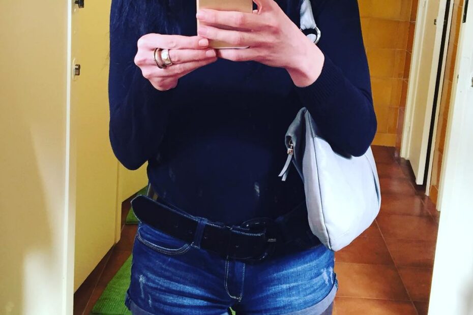 Selfie en el espejo de un lavabo: jersey oscuro, pantalón corto vaquero y bolso blanco