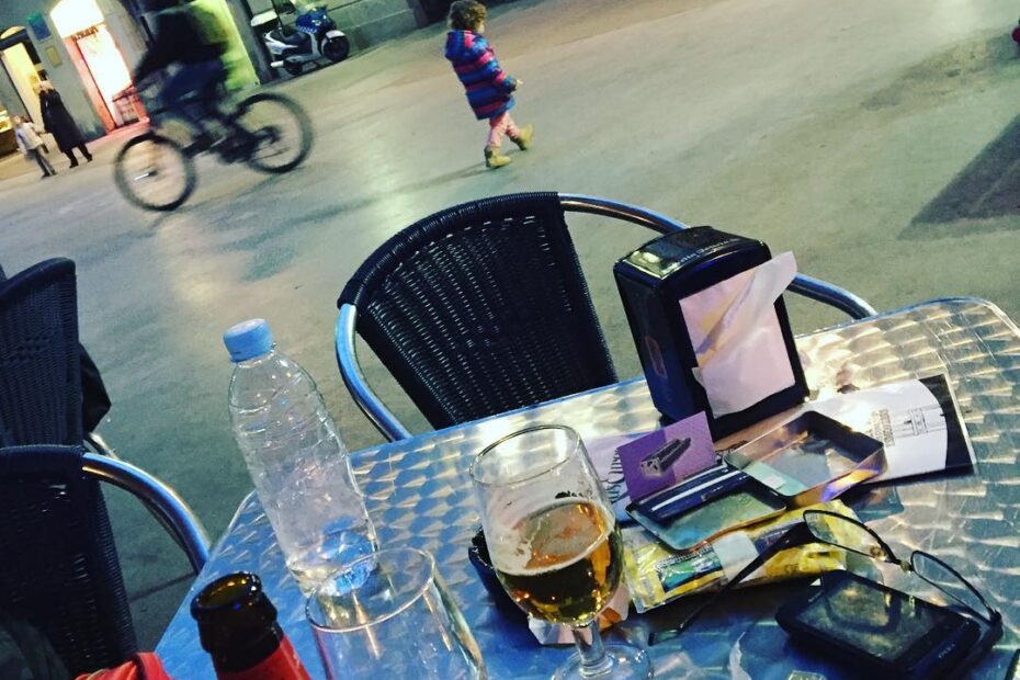Mesa de terraza con una botella de agua, una cerveza y objetos personales, ante una plaza con un ciclista y gente al fondo