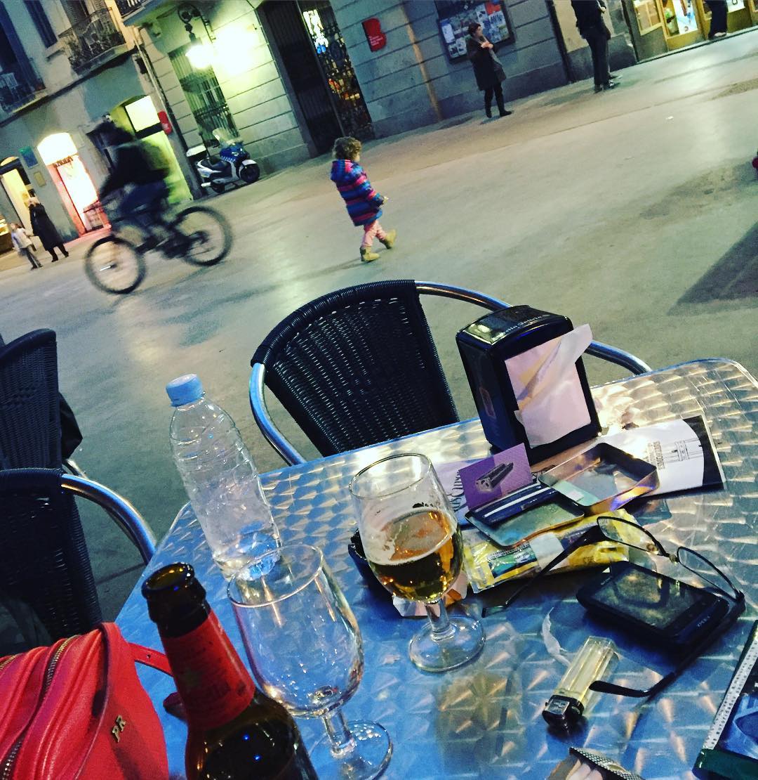 Mesa de terraza con una botella de agua, una cerveza y objetos personales, ante una plaza con un ciclista y gente al fondo