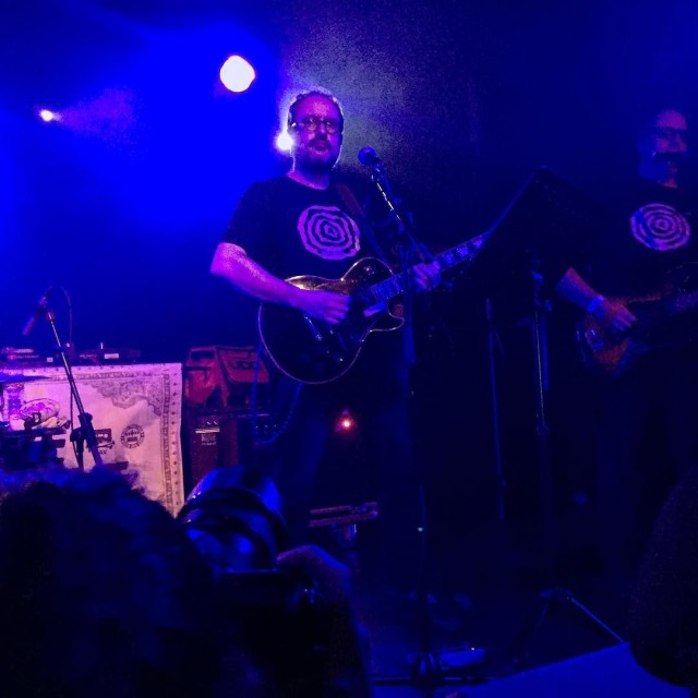 Guitarrista con camiseta negra actuando en un escenario con luz azul, en un concierto en la sala Apolo