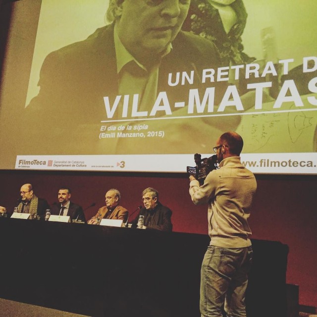 Quatre homes en una taula davant d'una pantalla amb el text «Un retrat de Vila-Matas» a la Filmoteca de Catalunya, amb un càmera gravant