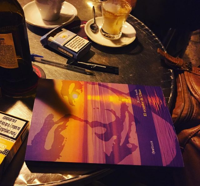 Un ejemplar de la novela El ecuador de Ulises, de Roser Amills, sobre la mesa de un bar con café, bolígrafos y una cajetilla de tabaco