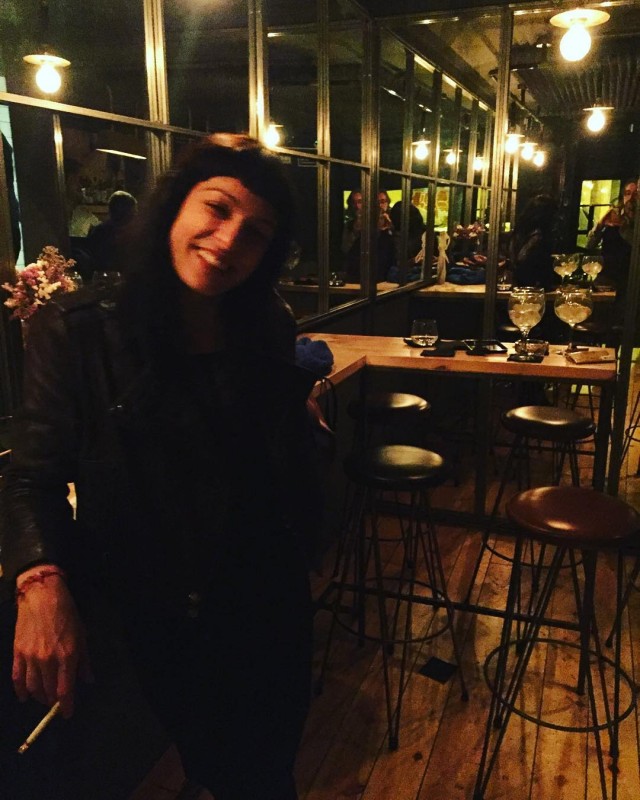 Mujer de pelo oscuro y flequillo, con chaqueta, de pie en un bar de noche con taburetes altos