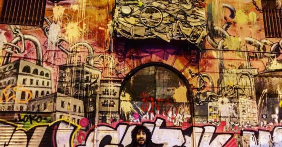 Hombre barbudo y de pelo rizado sentado delante de un mural de grafiti en una calle de Barcelona