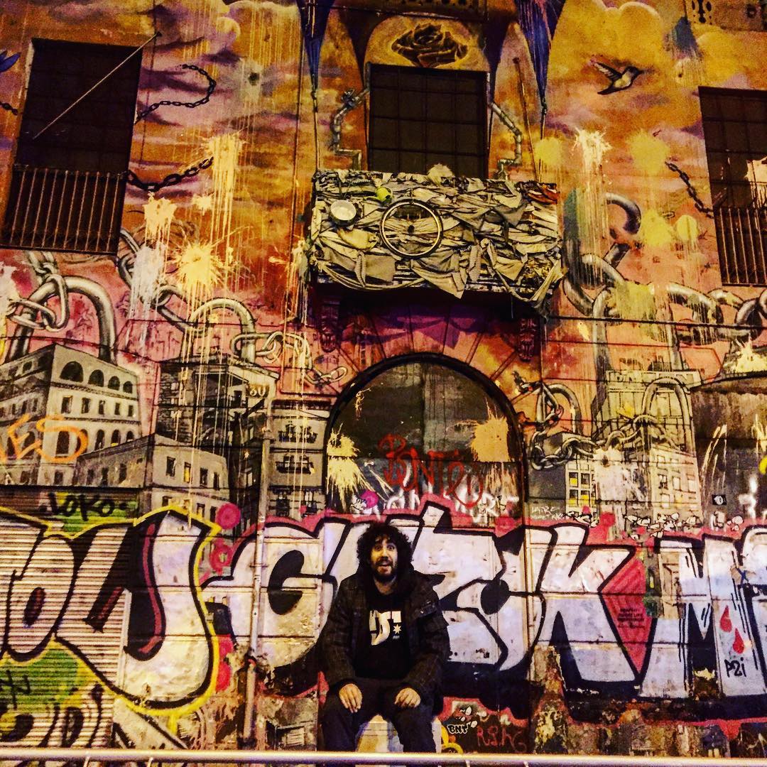 Hombre barbudo y de pelo rizado sentado delante de un mural de grafiti en una calle de Barcelona