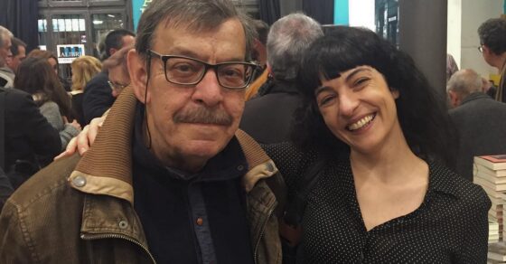Roser Amills sonriente junto al escritor José Luis Muñoz, con una copa de vino, entre el público de la librería Alibri Llibreria