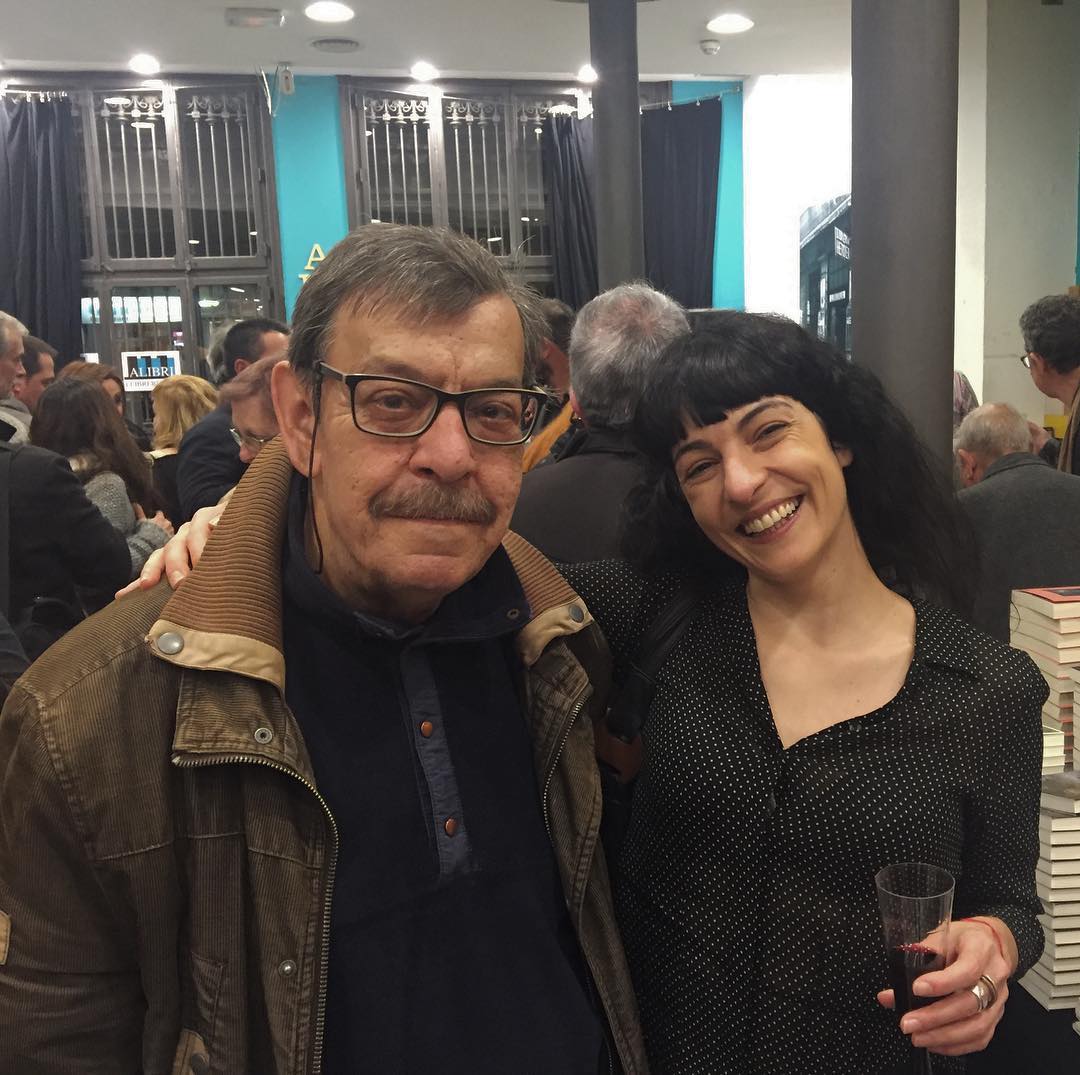 Roser Amills sonriente junto al escritor José Luis Muñoz, con una copa de vino, entre el público de la librería Alibri Llibreria
