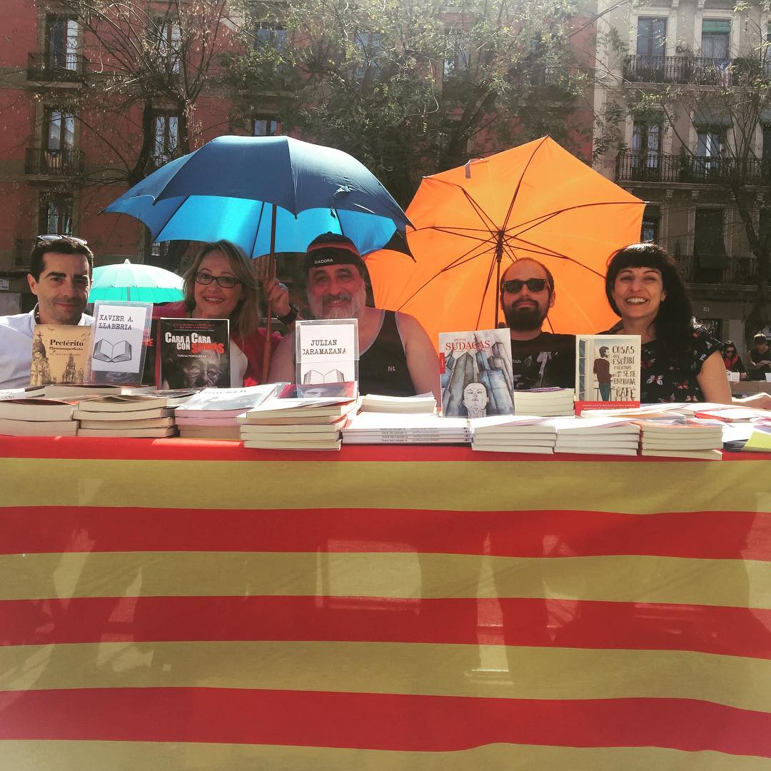 Roser Amills y otros autores firman libros en una mesa cubierta con una senyera, bajo paraguas azules y naranjas, en la plaça de la Vila de Gràcia