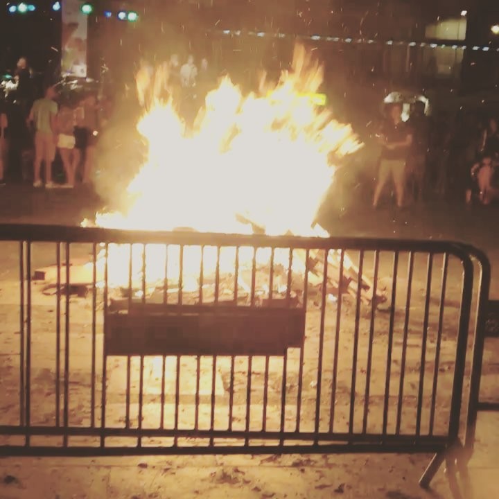 Una hoguera de Sant Joan ardiendo en plena calle tras una valla metálica, con gente alrededor