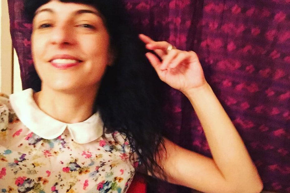 Roser Amills, con blusa de flores y cuello blanco, sonriente y con una mano levantada ante una cortina granate