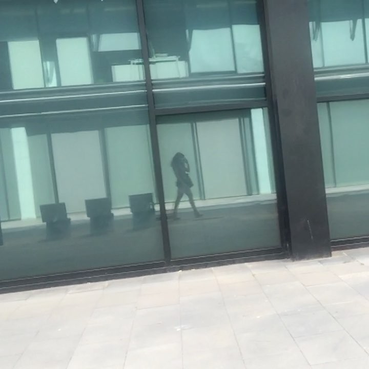El reflejo de Roser Amills caminando en la fachada de cristal oscuro de un edificio, sobre una acera