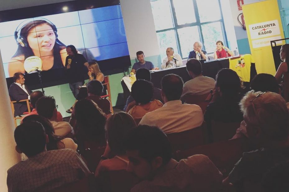 Taula del programa Revolució 4 amb els convidats i una pantalla gegant amb una dona amb auriculars, davant del públic de la Fàbrica Estrella Damm
