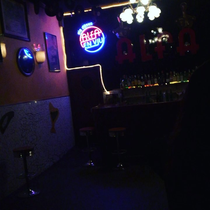Interior oscuro de un bar con un rótulo de neón azul y rojo con la palabra «Alfa» sobre la barra