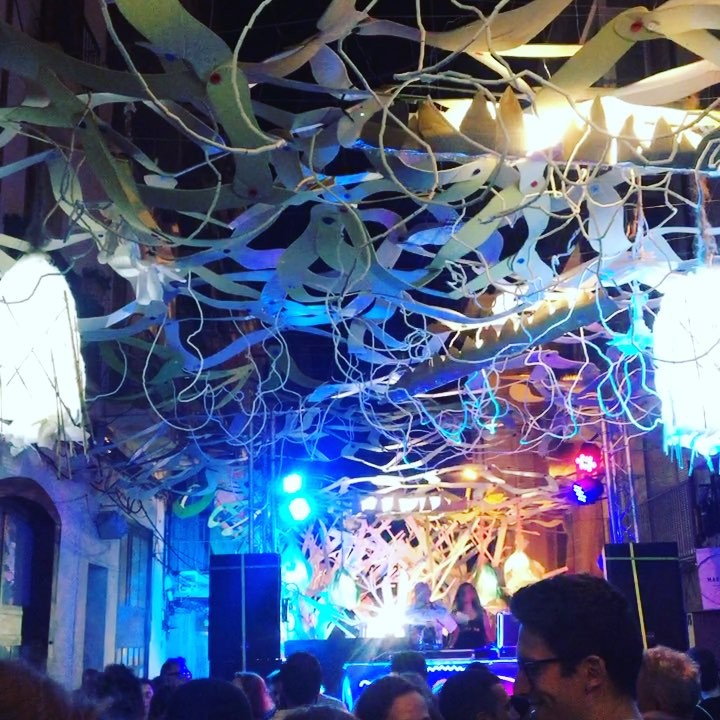 Calle de Gràcia engalanada con guirnaldas de papel y farolillos, con un escenario iluminado al fondo y público