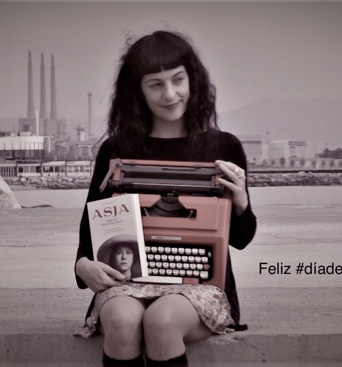Roser Amills sentada con una máquina de escribir y la novela «Asja», con chimeneas industriales al fondo