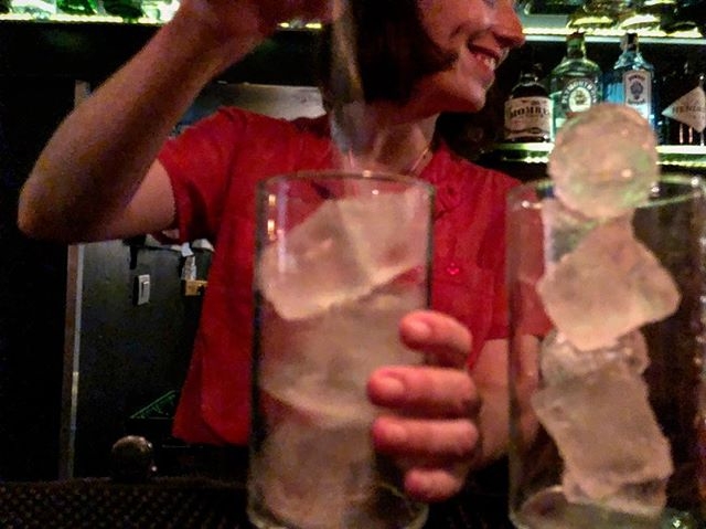 Una persona sirve bebida en dos vasos con grandes cubos de hielo, delante de una barra con botellas