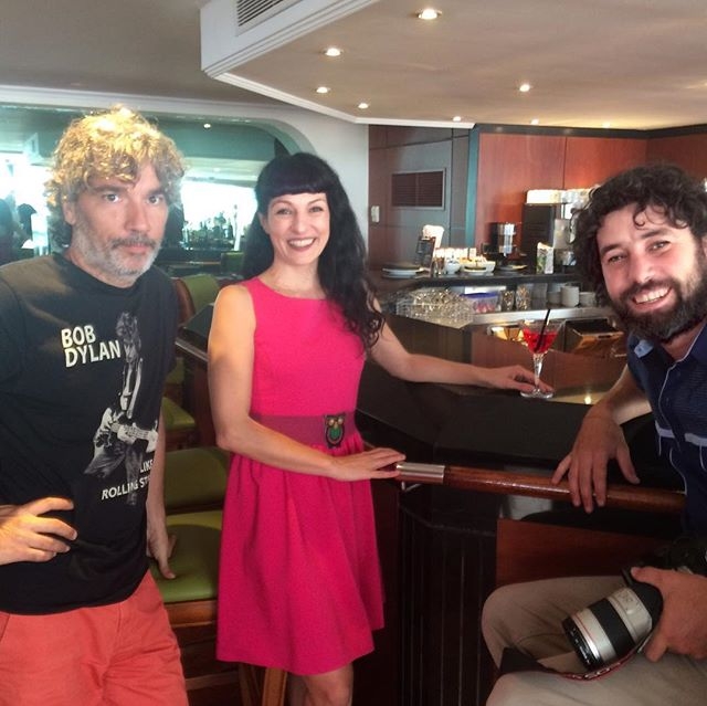 Tres personas posan junto a la barra de un local: un hombre con camiseta de Bob Dylan, una mujer con vestido rosa y una copa, y un hombre con barba y una cámara