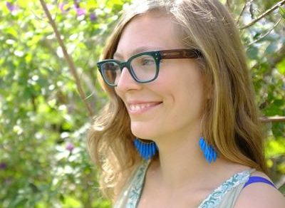 Retrato de Vanesa Berenstein con gafas y pendientes azules, entre vegetación