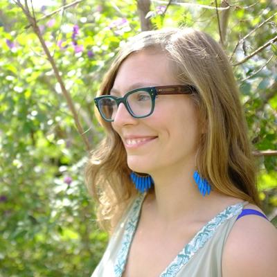 Retrato de Vanesa Berenstein con gafas y pendientes azules, entre vegetación