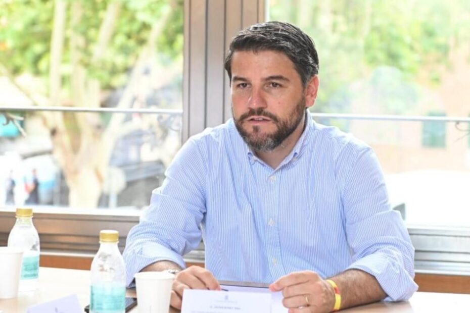 Home amb barba i camisa de ratlles blaves parlant assegut a una taula amb gots i ampolles d'aigua, davant d'una finestra