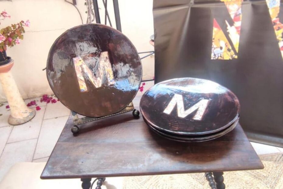 Dues peces de ceràmica fosca amb una gran «M» daurada, damunt una tauleta de fusta en un pati, juntament amb un cartell negre amb la lletra M