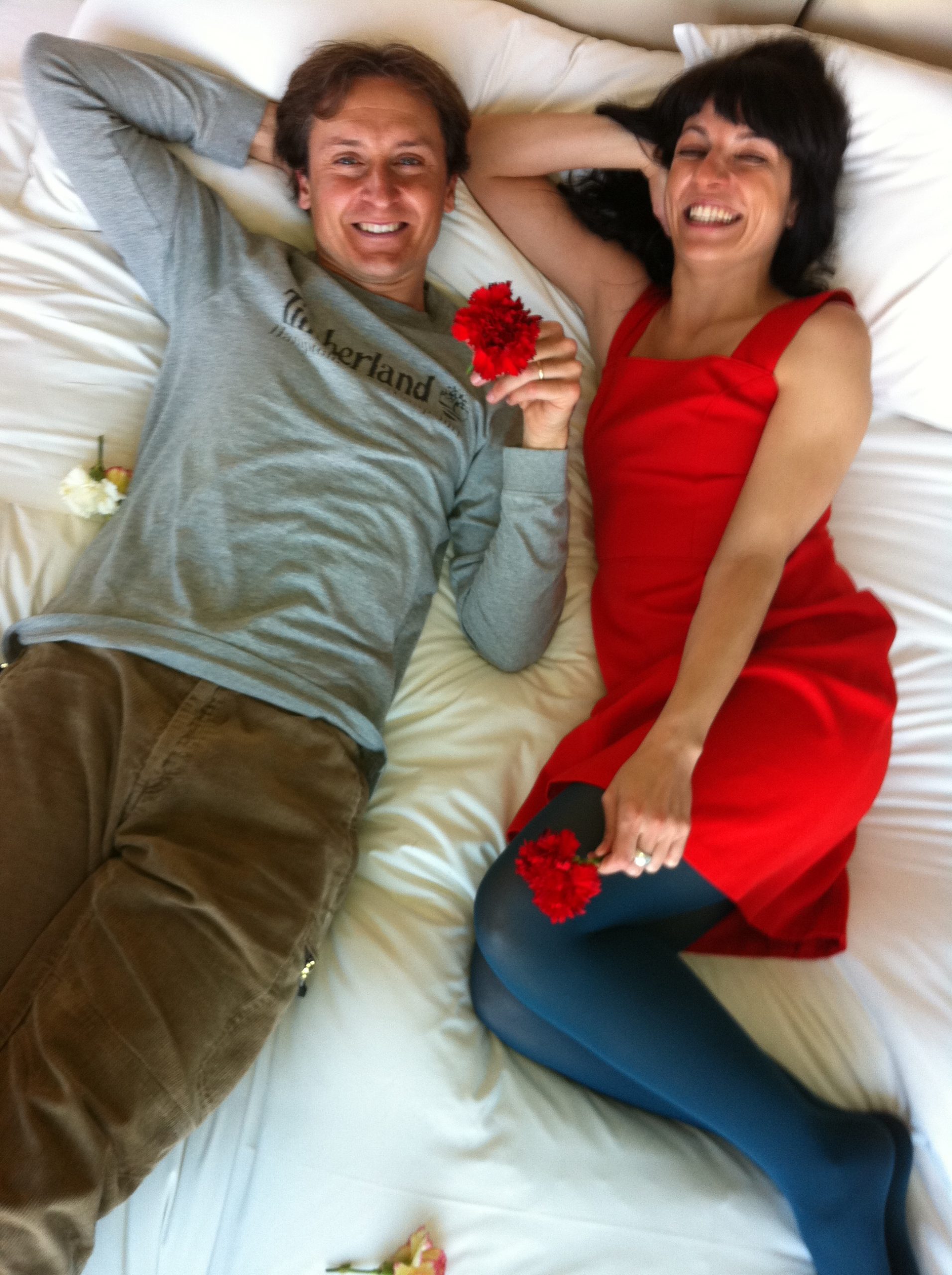 Roser Amills y Jordi Play tumbados sobre una cama blanca, ella con vestido rojo y rosas