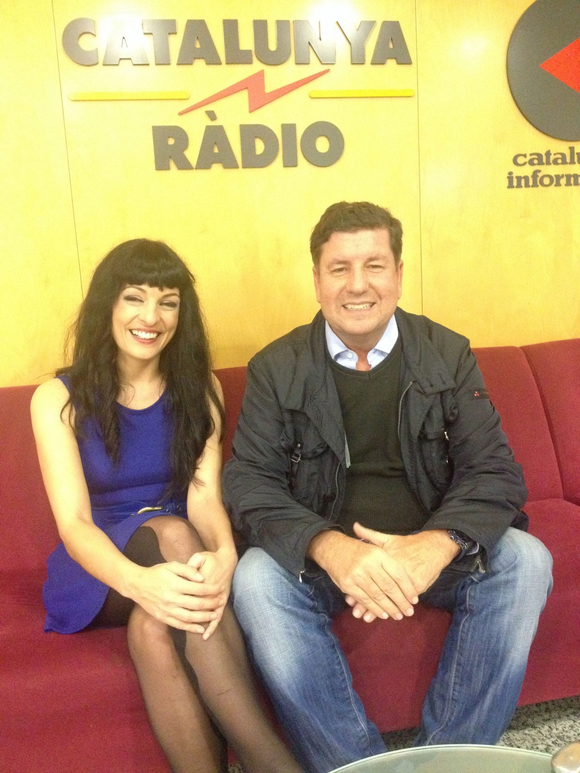 Roser Amills, con vestido azul, y Eduard Ruiz Castañé sentados en un sofá rojo ante el rótulo de Catalunya Ràdio