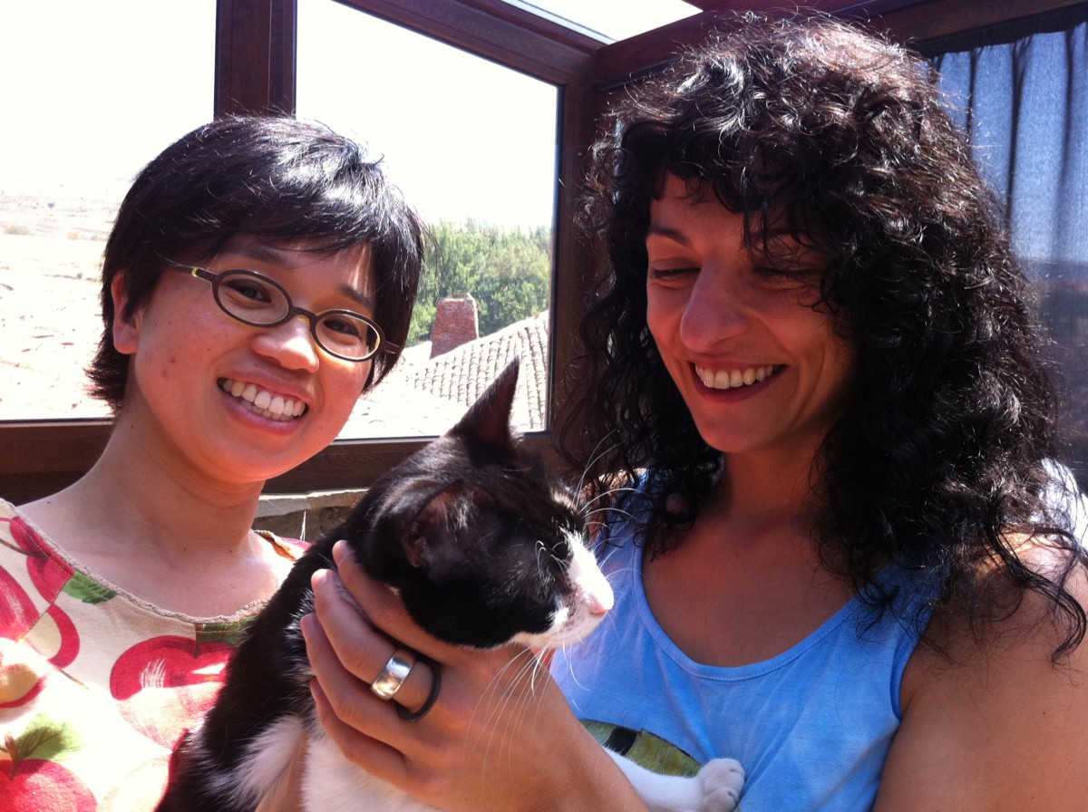 Naoko y Roser Amills ríen con un gato blanco y negro junto a una ventana con vistas a tejados