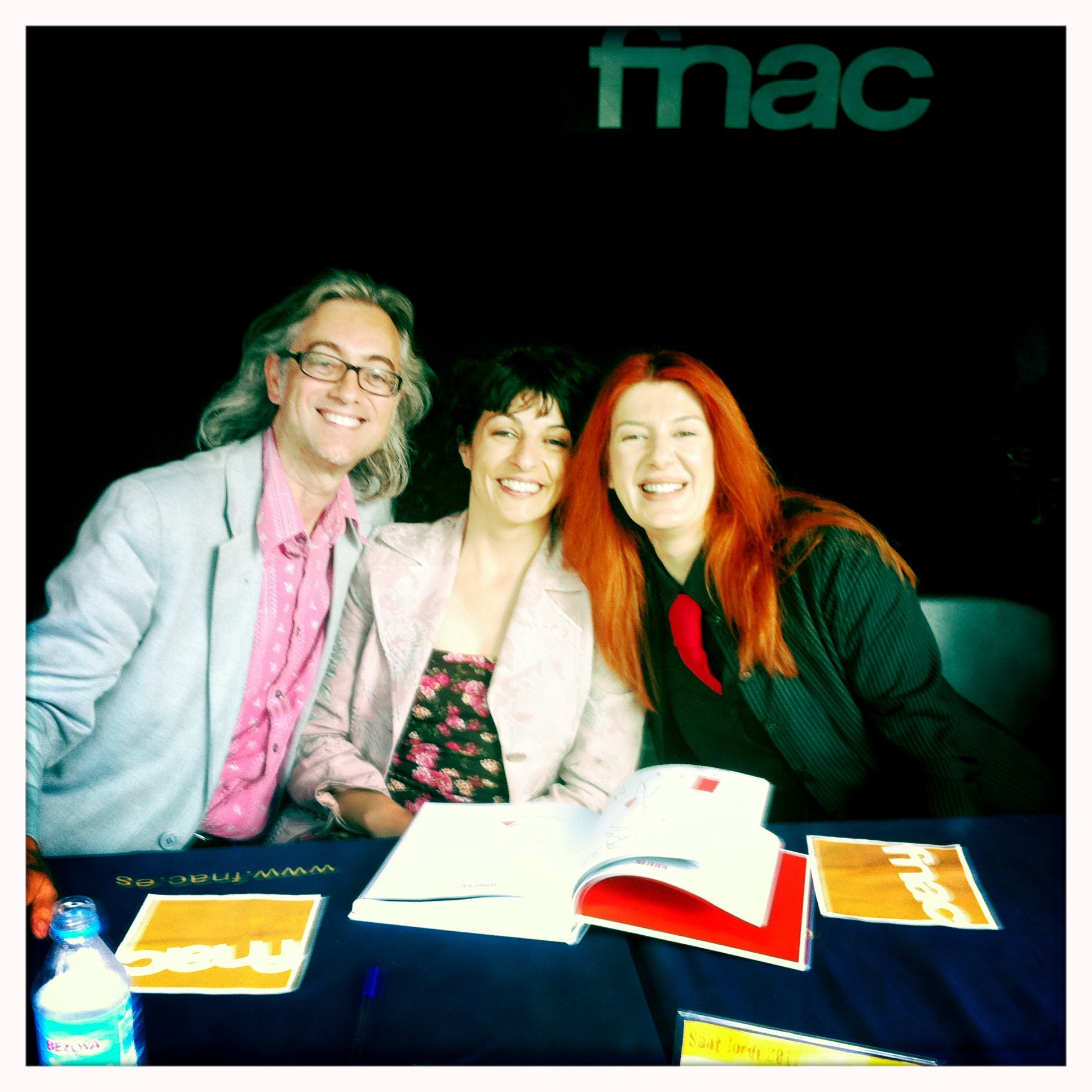 Tres persones assegudes en una taula de signatures de la FNAC, amb un llibre obert davant