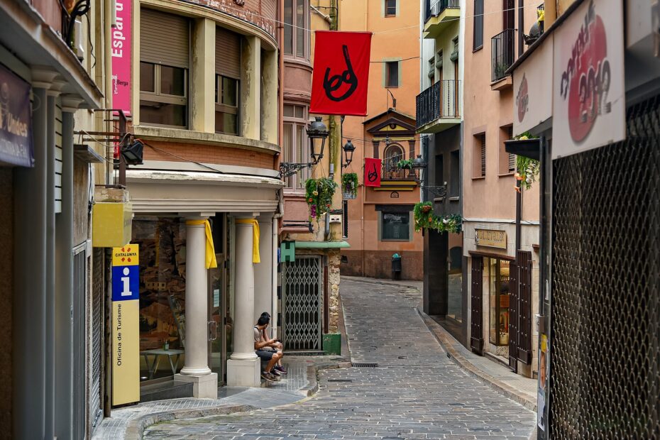 Calle empedrada y estrecha del centro histórico de Berga, con balcones, columnas a la entrada de un edificio y banderolas rojas colgadas
