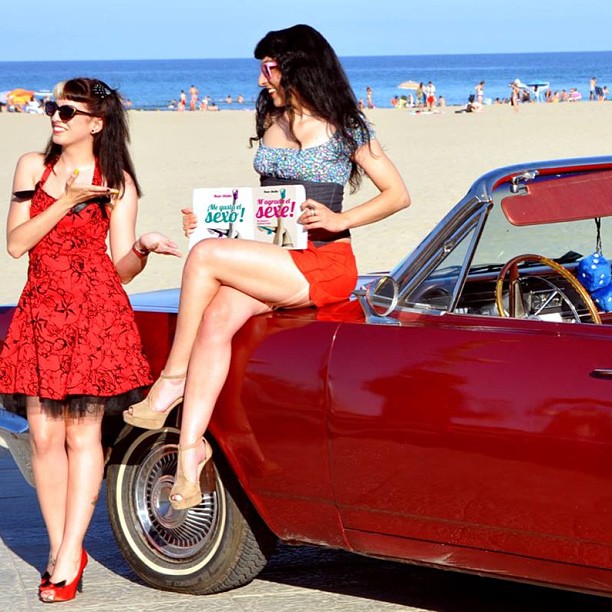 Roser Amills y una joven vestida de rockabilly junto a un coche descapotable rojo en la playa, con los libros Me gusta el sexo y M'agrada el sexe