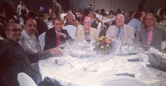 Siete hombres sentados a una mesa redonda con mantel blanco en la cena del Premio Planeta 2013; el de la derecha tiene el pelo largo y gafas